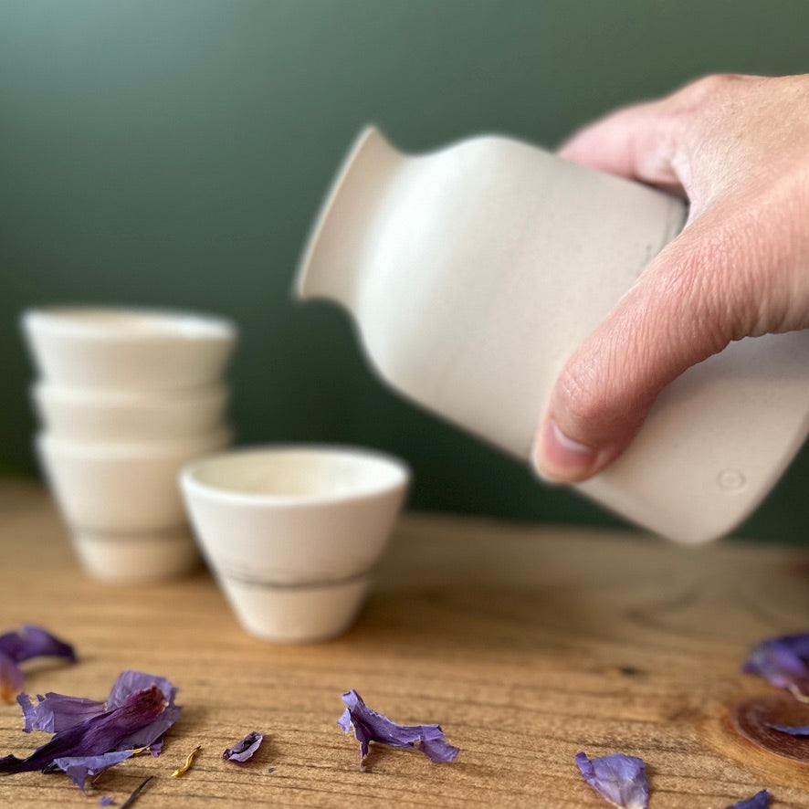 Person pouring from a white ceramic pitcher into small bowls on a wooden surface with purple petals.