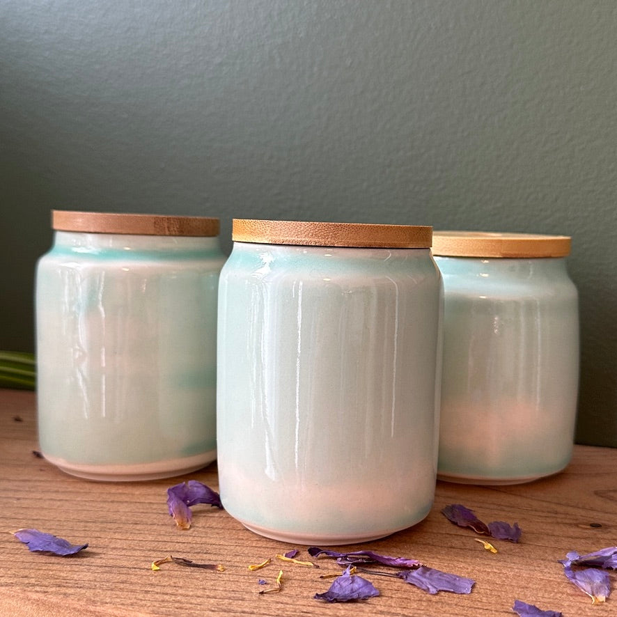 Three light blue ceramic jars with wooden lids on a wooden surface with purple flowers.