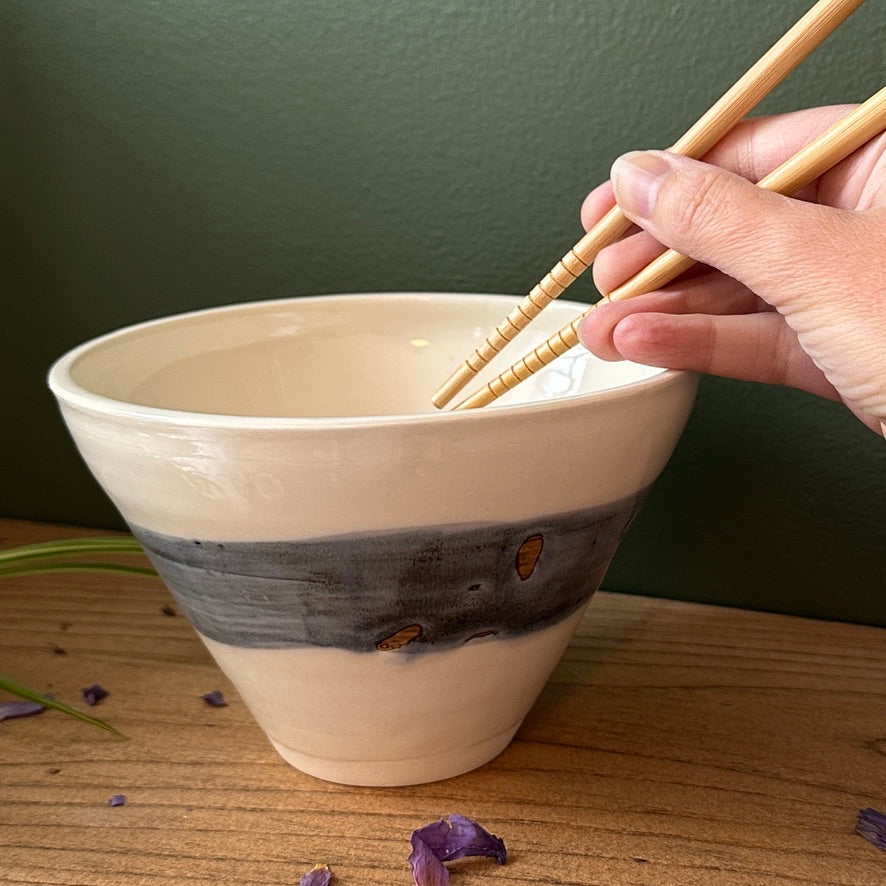 Hand holding chopsticks over a ceramic bowl on a wooden surface with a green background
