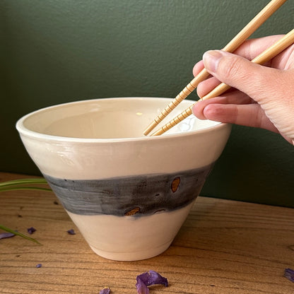 Hand holding chopsticks over a ceramic bowl on a wooden surface with a green background